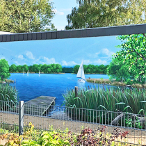 Ein See mit Segelboten, im Vordergrund ein Steg und Schilf daneben. Gesprüht auf eine Garagenseitenwand in Fürstenwalde von Mario Winkler.