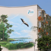 Ein Großes Wandbild mit einer Aussicht auf einen See, links mit Kiefern und es fliegt ein Storch am Himmel, gesprühtes Mural in Brandenburg, Storkow.