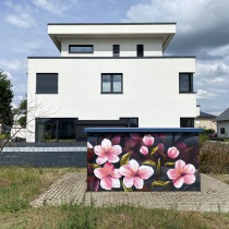 Ein Graffitiauftrag auf einer Trafostation mit Kirschblüten besprüht. Dahinter ein schönes neues Einfamilienhaus in Ludwigsfelde.